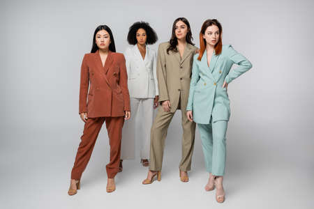 full length of multiethnic group of models in pastel color suits posing on gray