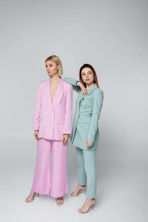 full length of blonde and redhead women in pink and blue pastel suits posing on gray