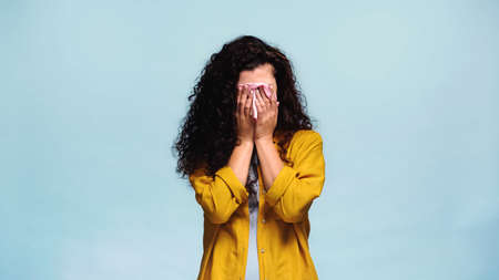 frustrated woman obscuring face with hands while crying isolated on blueの写真素材