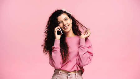 joyful woman touching hair and looking away while talking on smartphone isolated on pink