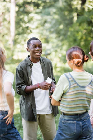 Positive african ameican teenager holding smartphone near friends on blurred foreground