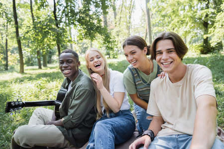 Happy teen girl sitting on grass near multiethnic friends with acoustic guitar