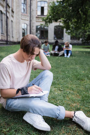 Student writing on notebook on lawn outdoors