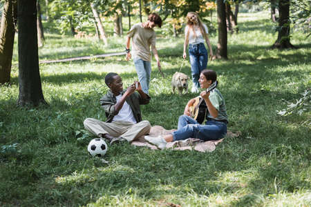 African american teenager using smartphone near friends playing acoustic guitar in parkの写真素材