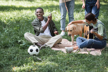 Cheerful african american boy holding smartphone near friends with retriever dog and acoustic guitar on lawnの写真素材