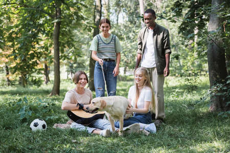 Smiling interracial teen friends spending time with retriever and acoustic guitar