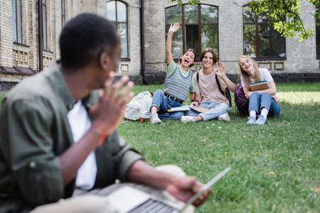 Cheerful students waving hands at blurred african american friend on lawnの写真素材