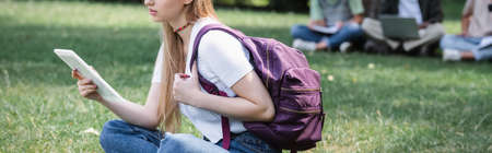 Cropped view of student holding backpack and digital tablet on grass, bannerの写真素材