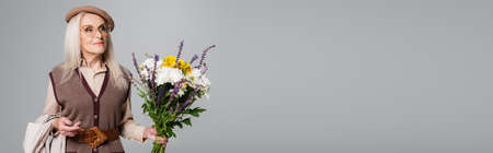 Elderly woman in beret holding flowers and trench coat isolated on gray, bannerの写真素材
