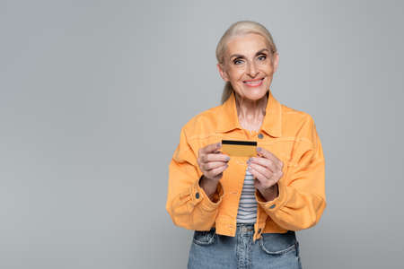 joyful senior woman holding credit card while looking at camera isolated on grayの写真素材