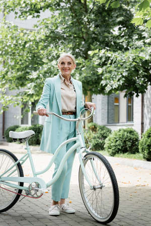 senior businesswoman in blue suit smiling near bicycle on urban streetの写真素材