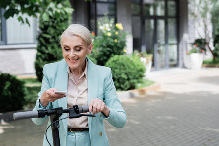 senior businesswoman smiling while messaging on smartphone near electric kick scooterの写真素材