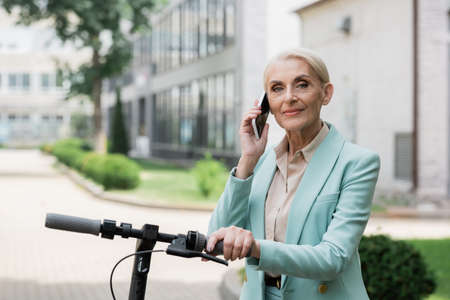 elderly businesswoman talking on mobile phone near electric kick scooter outdoorsの写真素材