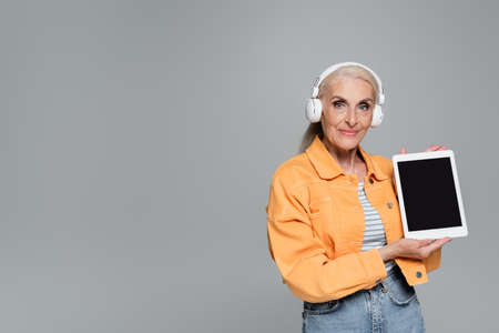 trendy senior woman in headphones showing digital tablet with blank screen isolated on gray