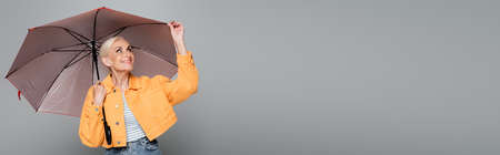 elderly woman in orange jacket adjusting umbrella isolated on gray, bannerの写真素材