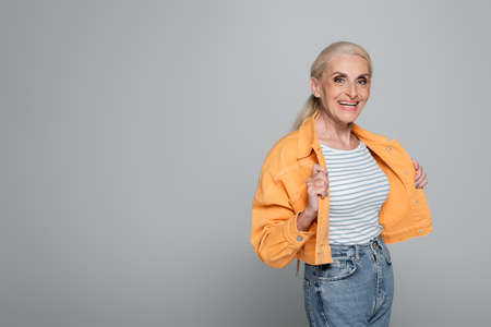 joyful senior woman in trendy jacket looking at camera while posing isolated on grayの写真素材