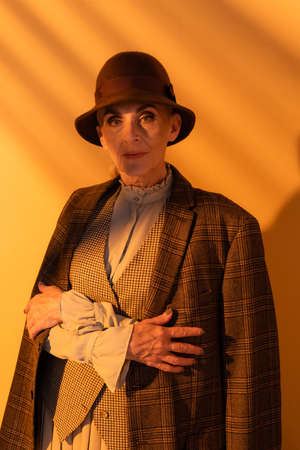Senior woman in hat and plaid jacket looking at camera on orange background
