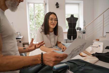 Asian designer looking at blurred colleague with sketch in studioの写真素材