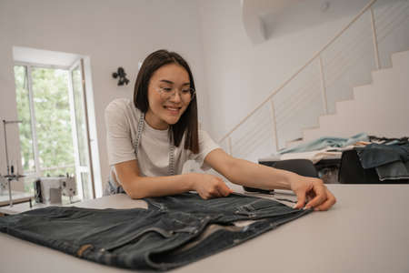 Cheerful asian designer in eyeglasses measuring jeans in studio