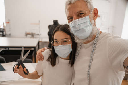 Designer in medical mask looking at camera near asian colleague with scissorsの写真素材