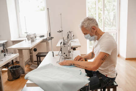 Tailor sewing medical mask near fabric in atelier