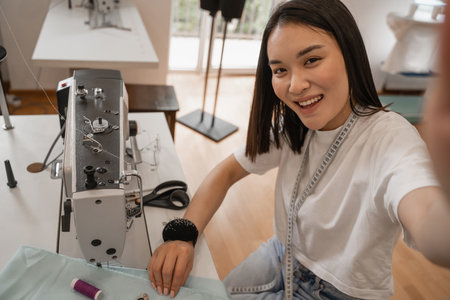 Smiling asian designer looking at camera near sewing machine and fabric