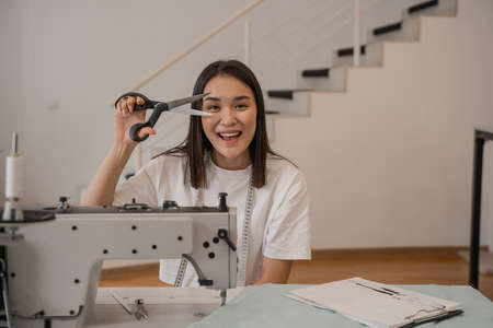 Cheerful asian designer holding scissors near sewing machine, fabric and clipboard