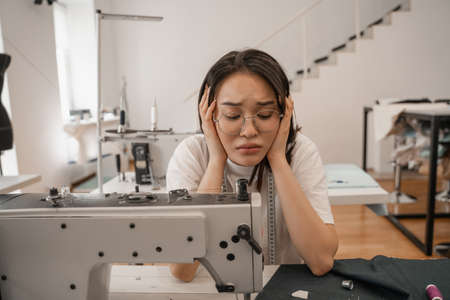 Tired asian seamstress looking at sewing machine and clothの写真素材