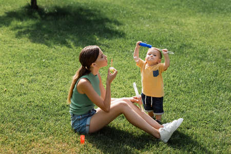 young mother sitting on grass and blowing soap bubbles near toddler son