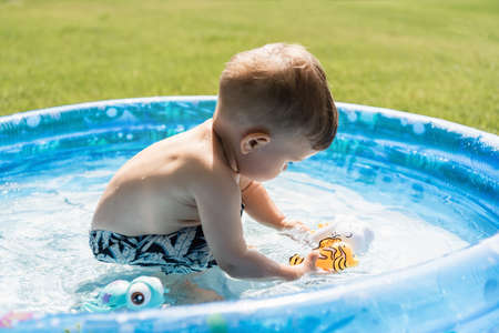 toddler boy in swim trunks playing with rubber toys in inflatable pool outside