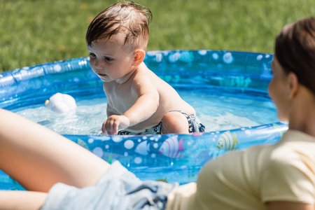 toddler boy sitting in inflatable pool near blurred mother on foreground