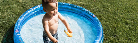 high angle view of cheerful toddler boy standing in inflatable pool with rubber toy, banner