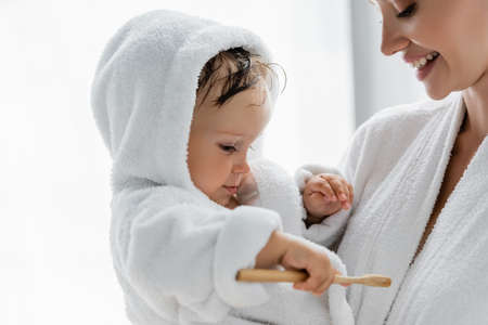 toddler boy in bathrobe holding toothbrush near happy motherの写真素材