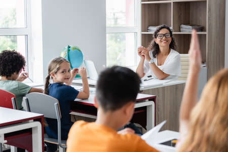 blurred schoolgirl raising hand near african american teacher and classmatesの写真素材