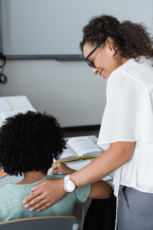 smiling african american teacher touching back of schoolboy in classroomの写真素材