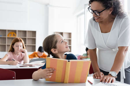 smiling schoolgirl and african american teacher looking at each other near blurred pupilsの写真素材