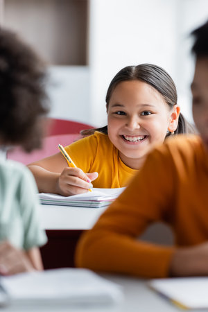 asian schoolgirl holding pen while smiling at camera near blurred boysの写真素材