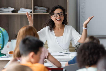 excited african american teacher showing wow gesture near blurred children in classroomの写真素材