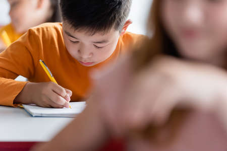 asian boy writing in notebook near blurred classmatesの写真素材