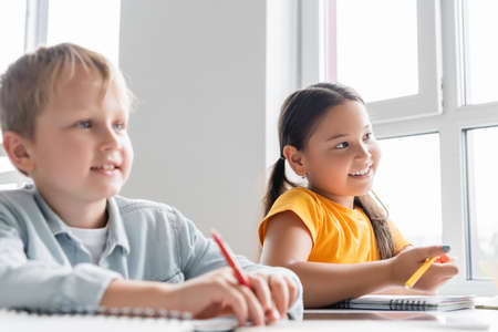 happy interracial schoolkids holding pens during lesson in classroom