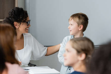 positive african american teacher talking to schoolboy during lesson near blurred pupilsの写真素材