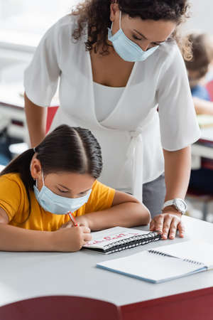 african american teacher in medical mask standing near asian girl writing in notebook