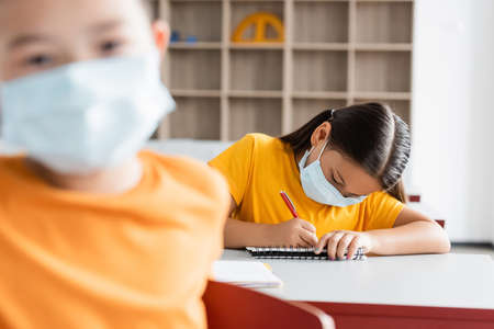 asian schoolgirl in medical mask writing in notebook during lessonの写真素材