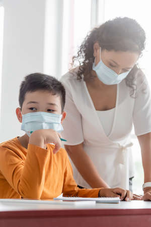 african american teacher in medical mask standing near asian schoolboy in classroomの写真素材