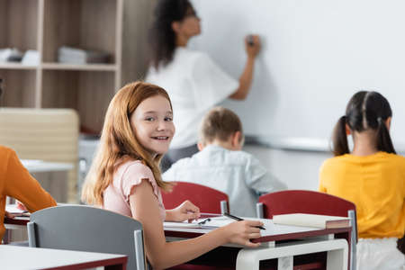 joyful redhead schoolgirl smiling at camera while blurred african american teacher writing on whiteboardの写真素材