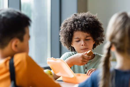 african american boy eating sandwiches near blurred classmates