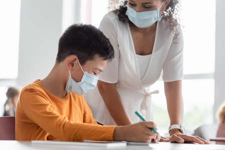 african american teacher in medical mask standing near asian boy in classroomの写真素材