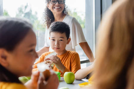 asian boy with sandwich near blurred girls and smiling african american teacherの写真素材