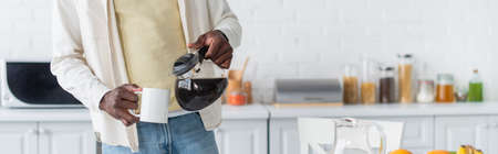 cropped view of african american man holding coffee pot near cup in kitchen, bannerの写真素材