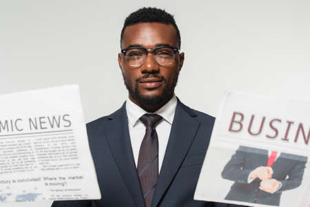 african american man in eyeglasses holding newspapers isolated on gray